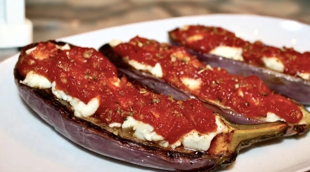 Przekrojony wzdłuż na połowę bakłażan pieczony, a następnie po obłożeniu serem feta i aromatycznym sosem pomidorowym i krótko zapiekany.