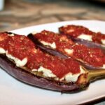 Przekrojony wzdłuż na połowę bakłażan pieczony, a następnie po obłożeniu serem feta i aromatycznym sosem pomidorowym i krótko zapiekany.