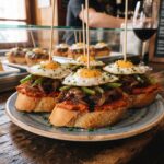Pincho czyli hiszpańska przekąska to kanapka posmarowana miękką kiełbasą paprykowaną, obłożona karmelizowaną cebulą, kawałkami papryki i wzbogacona sadzonymi jajkami przepiórczymi.