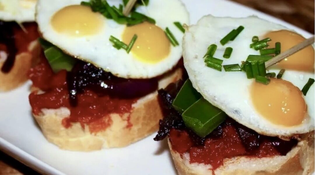 Pincho czyli hiszpańska przekąska to kanapka posmarowana miękką kiełbasą paprykowaną, obłożona karmelizowaną cebulą, kawałkami papryki i wzbogacona sadzonymi jajkami przepiórczymi.