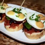 Pincho czyli hiszpańska przekąska to kanapka posmarowana miękką kiełbasą paprykowaną, obłożona karmelizowaną cebulą, kawałkami papryki i wzbogacona sadzonymi jajkami przepiórczymi.