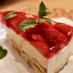 Sernik na zimno na spodzie z biszkoptów pokrytych masą z jajek, cukru i twarogu pokrytej galaretką i ozdobioną truskawkami.