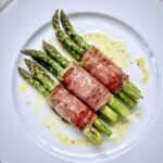 Zielone szparagi owinięte plastrami szynki dojrzewającej, pieczone i skropione oliwą extra vergine tuż przed podaniem.