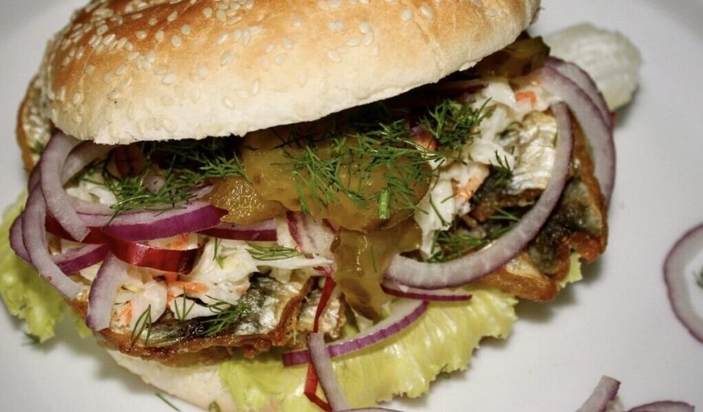 Pyszny burger ze śledziem smażonym podanym w bułce razem z dodatkiem sałatki Coleslaw, ogórkiem kiszonym i czerwoną cebulą.