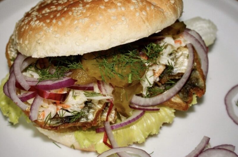 Pyszny burger ze śledziem smażonym podanym w bułce razem z dodatkiem sałatki Coleslaw, ogórkiem kiszonym i czerwoną cebulą.