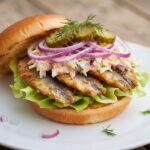 Pyszny burger ze śledziem smażonym podanym w bułce razem z dodatkiem sałatki Coleslaw, ogórkiem kiszonym i czerwoną cebulą.