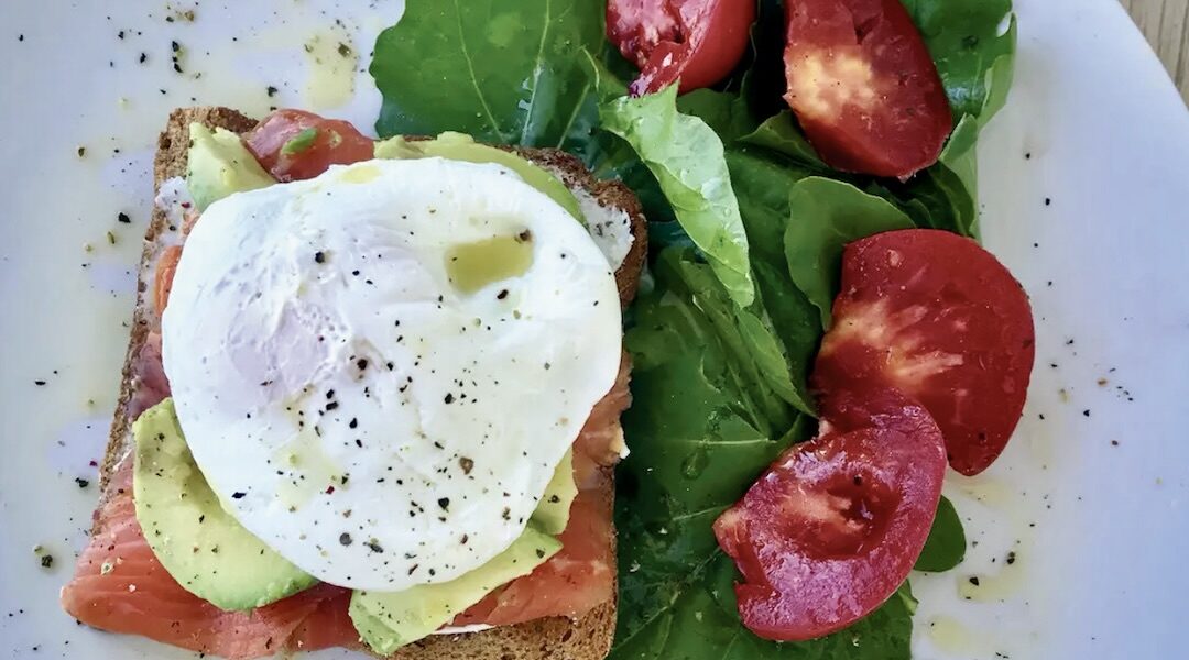 Kanapka z łososiem wędzonym na zimno przygotowana z chleba tostowego posmarowanego serkiem obłożona dodatkowo awokado i jajkiem w koszulce.