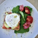 Kanapka z łososiem wędzonym na zimno przygotowana z chleba tostowego posmarowanego serkiem obłożona dodatkowo awokado i jajkiem w koszulce.