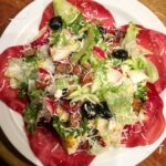 Sałatka z cieniutko pokrojoną w plasterki suszoną wołowiną bresaolą podana z miksem sałat, oliwkami, jajkiem i rzodkiewkami w sosie winegret.