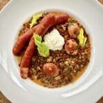 Soczewica gotowana z krojonymi warzywami doprawiona śmietaną i wzbogacona smażoną kiełbasą wędzoną w plastrach.