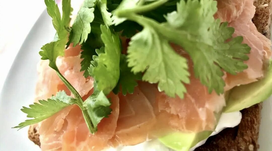 Tosty posmarowane serkiem i obłożone wędzonym łososiem z dodatkiem awokado i listkami kolendry.