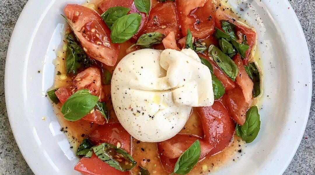 Burrata to unikatowy delikatny ser mozzarellą wypełniony śmietanką podany w tym przepisie razem z pomidorami koktajlowymi oliwa i świeżą bazylią.