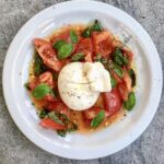 Burrata to unikatowy delikatny ser mozzarellą wypełniony śmietanką podany w tym przepisie razem z pomidorami koktajlowymi oliwa i świeżą bazylią.