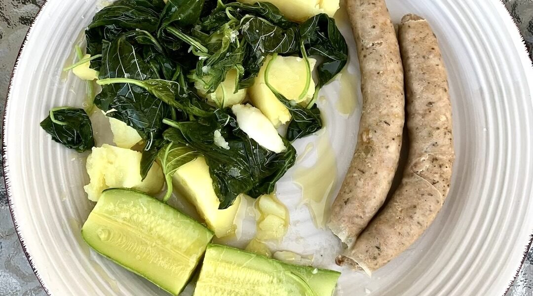 Białe kiełbaski gotowane i podane z warzywami gotowanym, a konkretnie z czosnkiem, ziemniakami, szpinakiem i cukinią, polanymi przed podaniem oliwą i sokiem z cytryny.