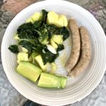 Białe kiełbaski gotowane i podane z warzywami gotowanym, a konkretnie z czosnkiem, ziemniakami, szpinakiem i cukinią, polanymi przed podaniem oliwą i sokiem z cytryny.