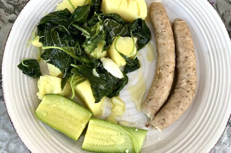 Białe kiełbaski gotowane i podane z warzywami gotowanym, a konkretnie z czosnkiem, ziemniakami, szpinakiem i cukinią, polanymi przed podaniem oliwą i sokiem z cytryny.