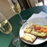 na zielonym stole znajduje się biały talerzyk z kanapkami, a obok stoją dwa kieliszki z szampanem