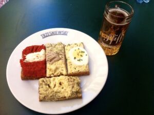 na stole znajduje się biały talerz z czterema kanapkami, a obok talerza kufelek z piwem