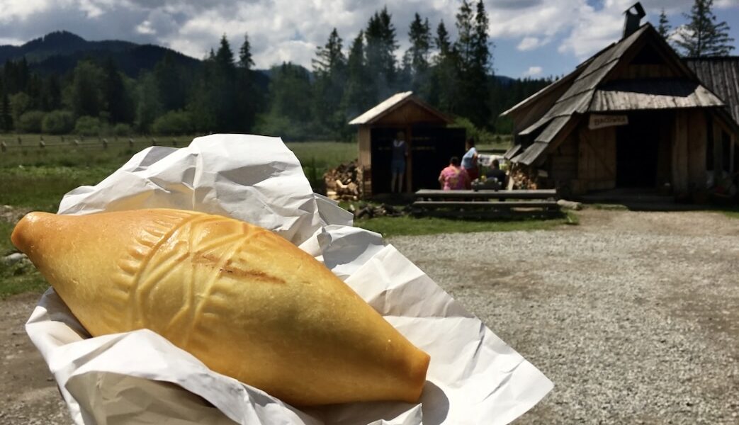 świeżo wędzony oscypek na papierze trzymany w dłoni z bacówką w tle