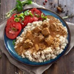 gulasz z łopatki cielęcej na talerzu podany na kaszy jęczmiennej w towarzystwie kilku plastrów pomidora posypanych siekaną dymką