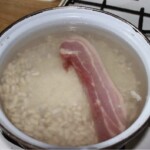 w dużym garnku znajduje się biała fasolka oraz duży plaster boczku wędzonego