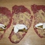 trzy plastry wołowiny na rolady śląski posmarowane musztardą i obłożone piórkami cebuli i słupkami ogórka kiszonego