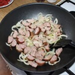 na patelni smażą się kawałki kiełbasy razem z krojoną cebulą